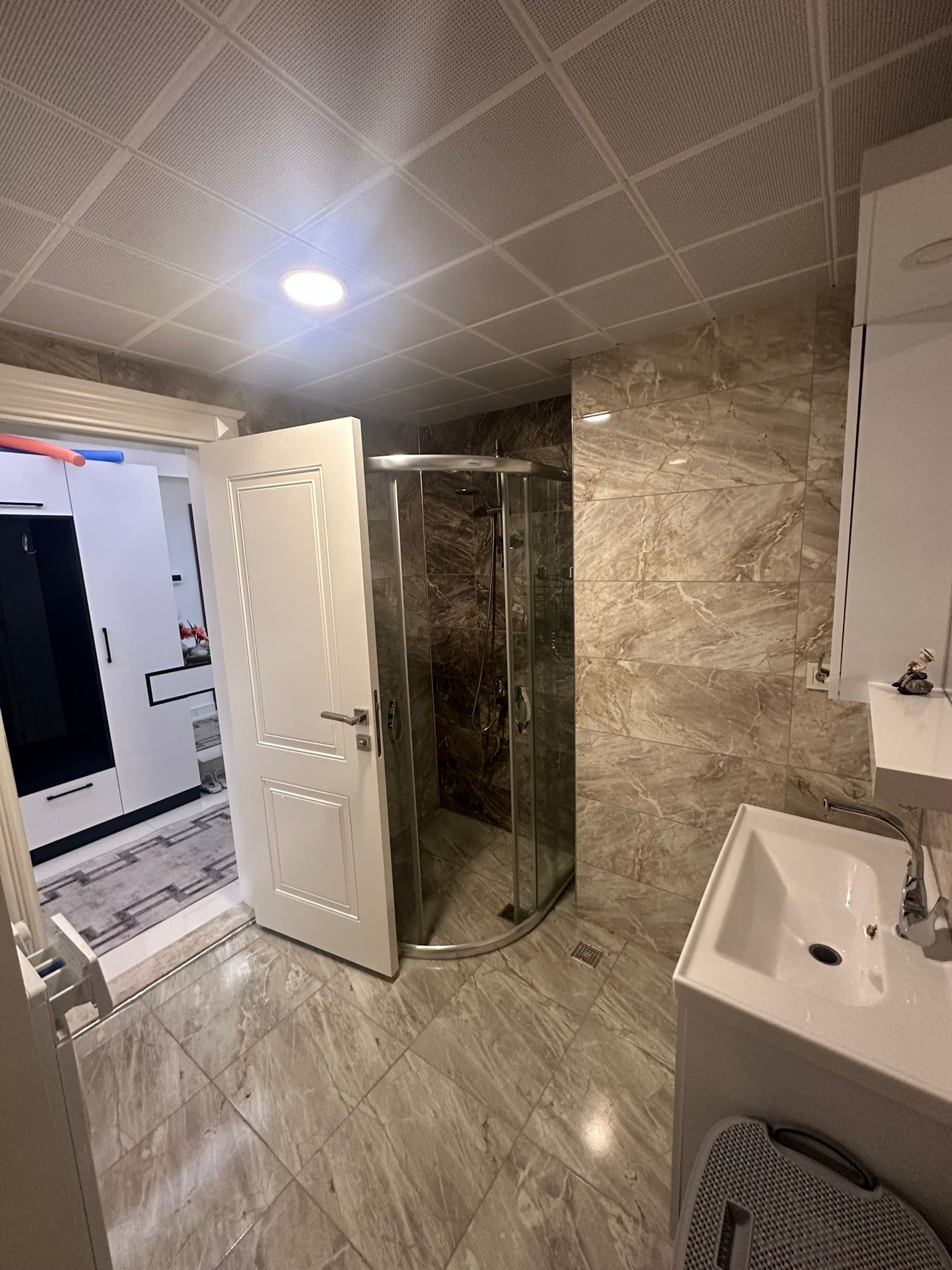 Alanya’daki konutun banyosunda yarım daire cam duş kabini, beyaz lavabo ve mermer fayans
