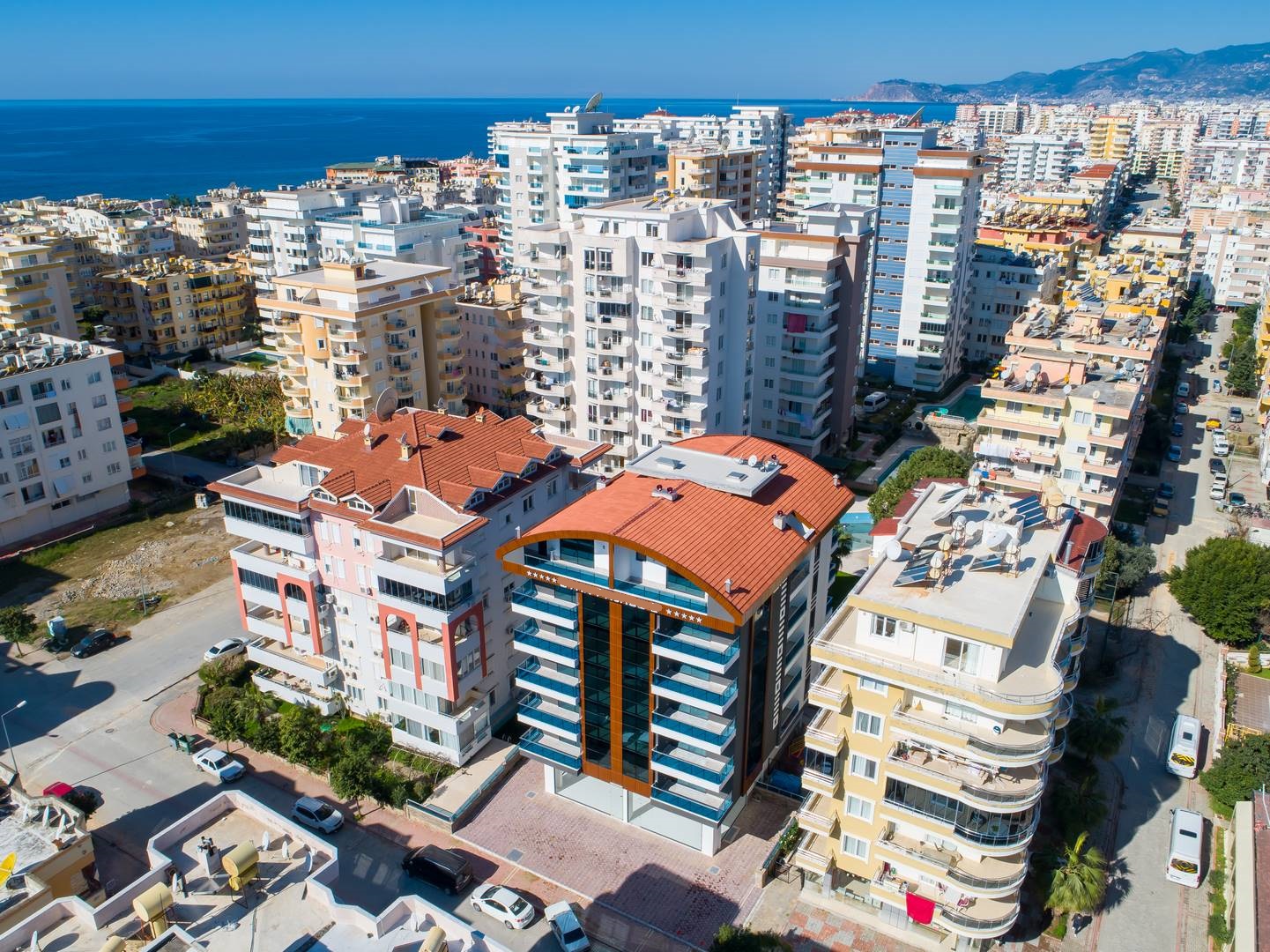Kıvrımlı kırmızı çatılı bina, Alanya’da satılık konutların yer aldığı sokaklarda uzaktaki deniz kıyısıyla görülüyor