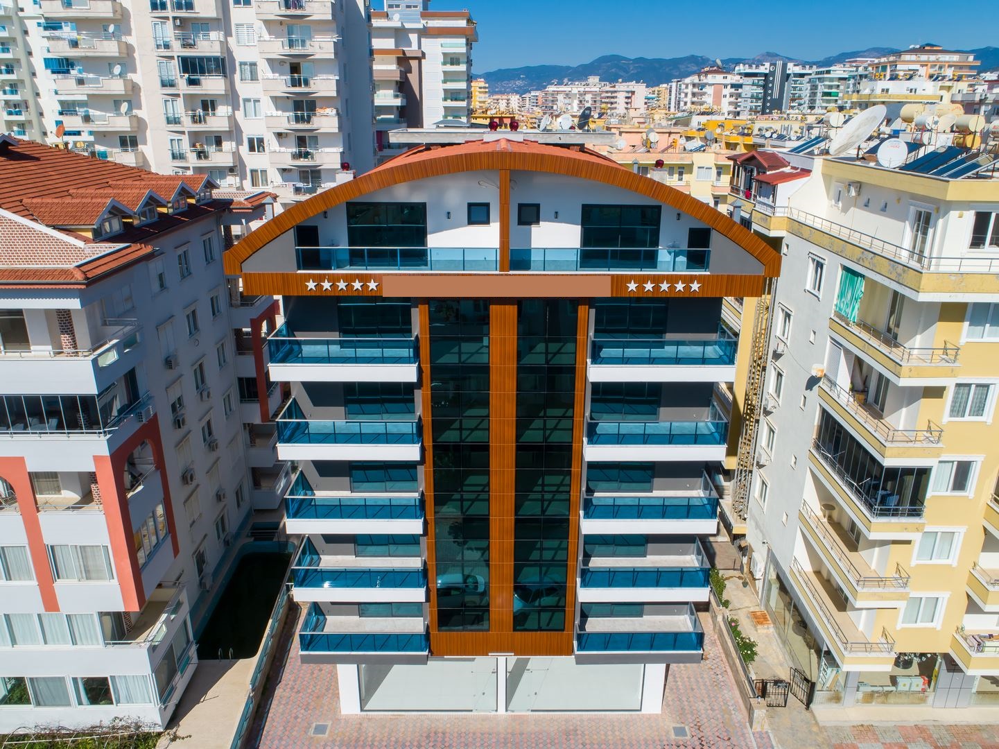 Simetrik cepheli daire binasında dalgalı kırmızı çatı ve iki sıra mavi cam balkon, Alanya’da