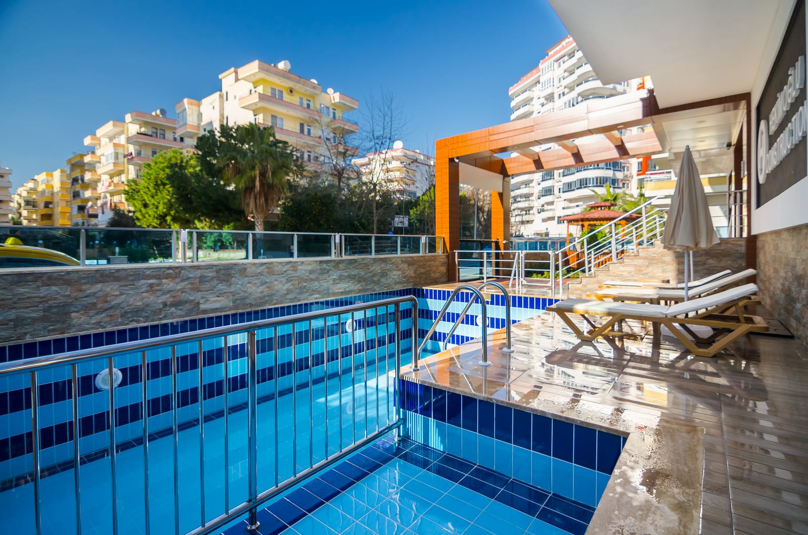 Açık havuzda metal basamak korkulukları, ahşap şezlonglar ve geçit üzerindeki pergola