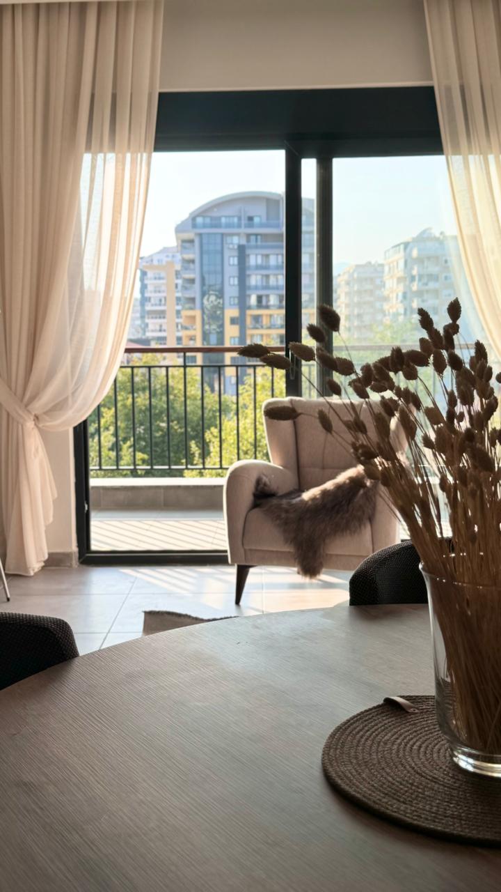 Yemek alanında yuvarlak ahşap masa, kuru çiçekli vazo ve denize yakın balkona açılan cam kapı