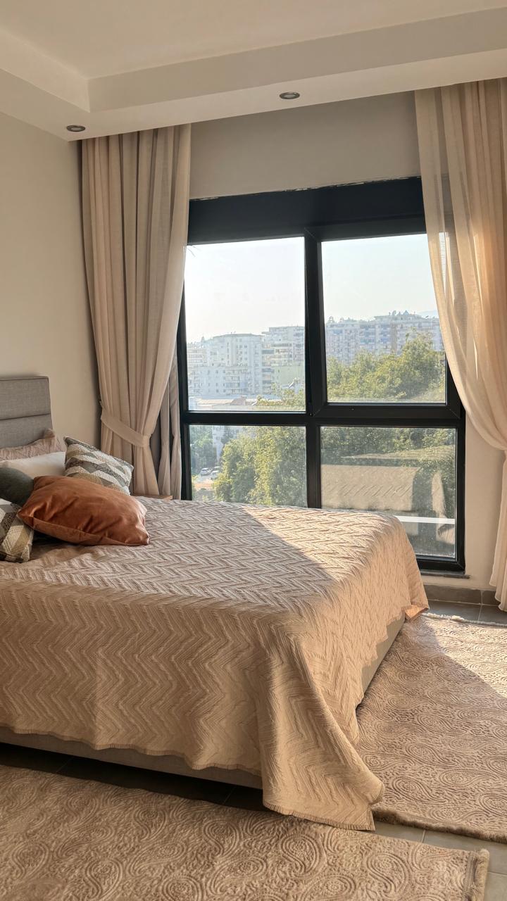 Panoramik pencereli yatak odasında bej örtülü çift kişilik yatak ve kentteki evler manzarası