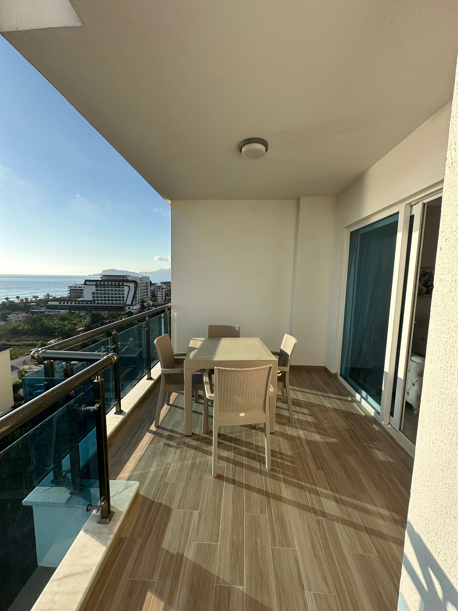 Satılık dairenin uzun kapalı balkonunda rattan yemek takımı ve panoramik akşam deniz manzarası