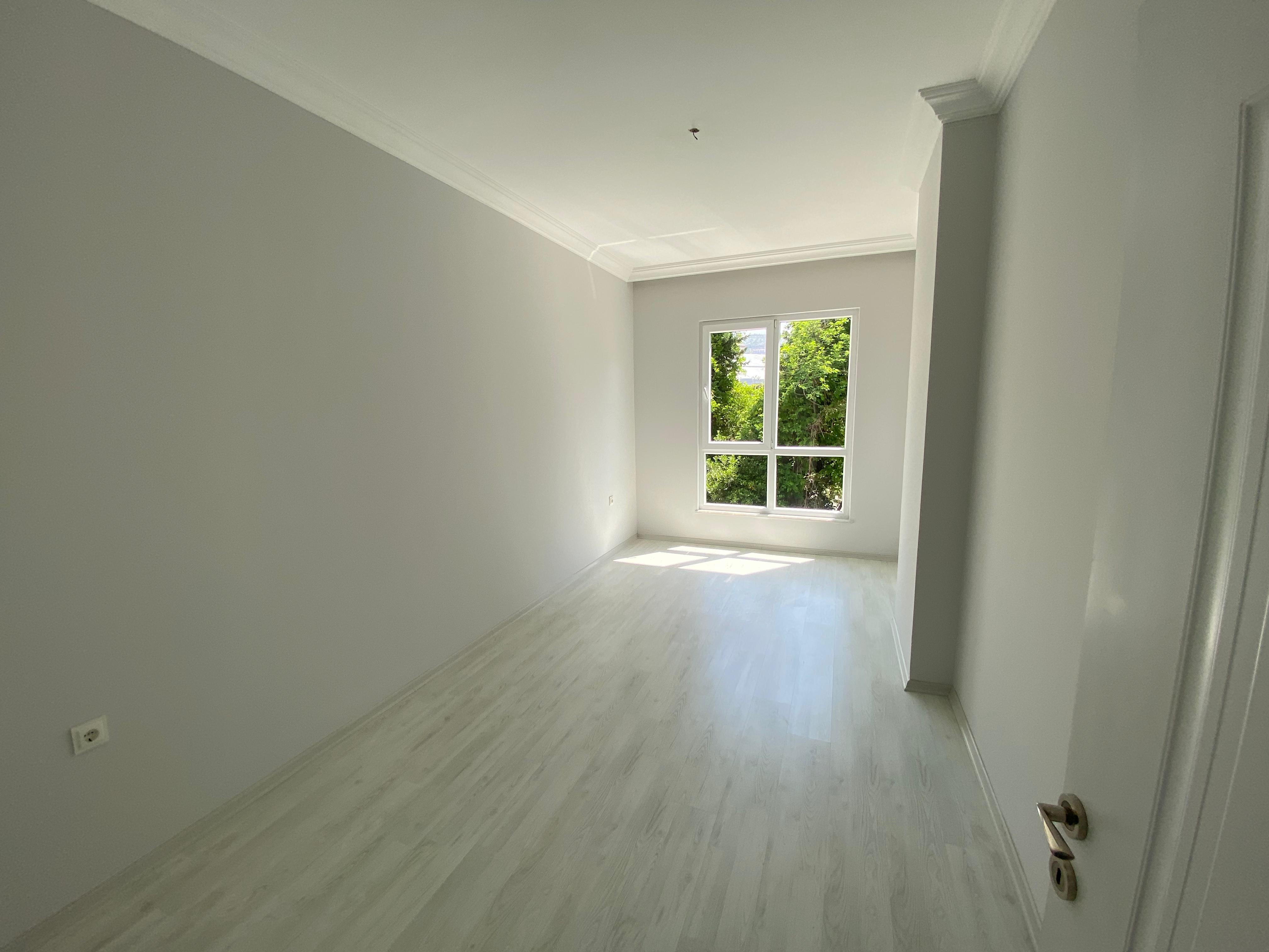 2+1 dairenin aydınlık odasında gri duvarlar, açık renk laminat ve ağaçlara bakan çift kanatlı pencere