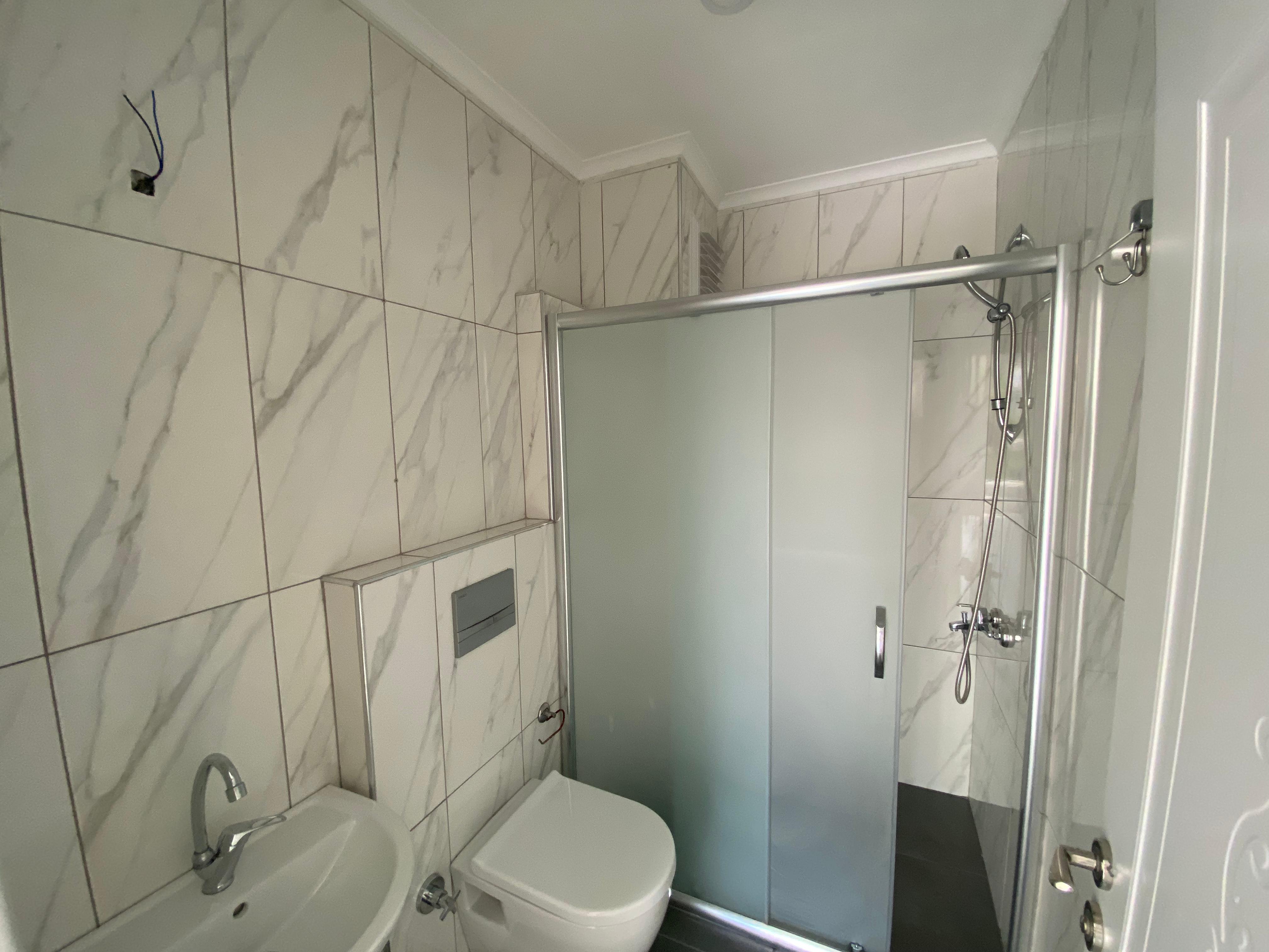 Alanya’daki 2+1 evin banyosunda buzlu cam duş kabini, beyaz mermer görünümlü duvarlar ve lavabo