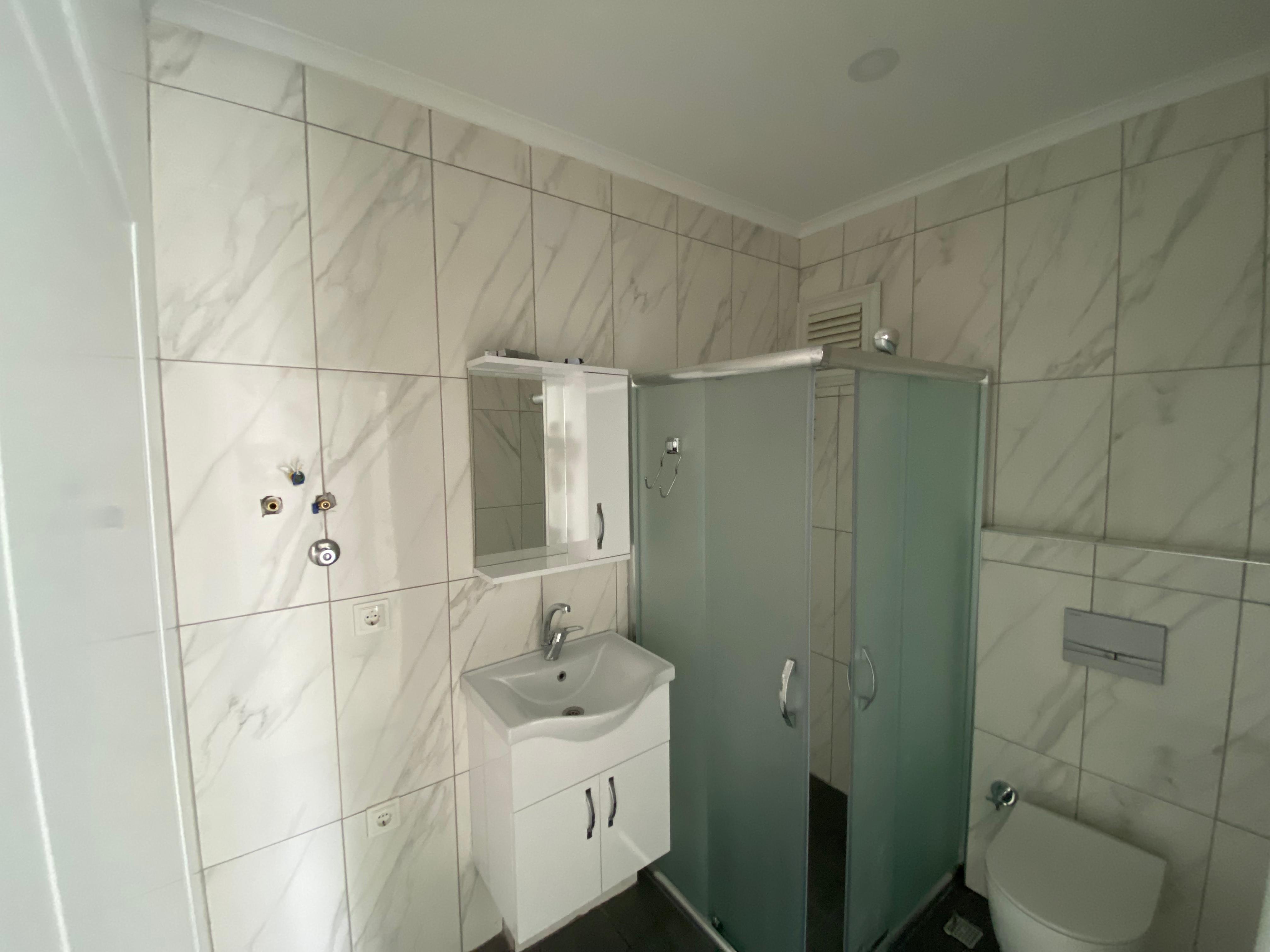 Banyoda buzlu cam duş kabini, aynalı lavabo dolabı ve koyu renk zemin karoları