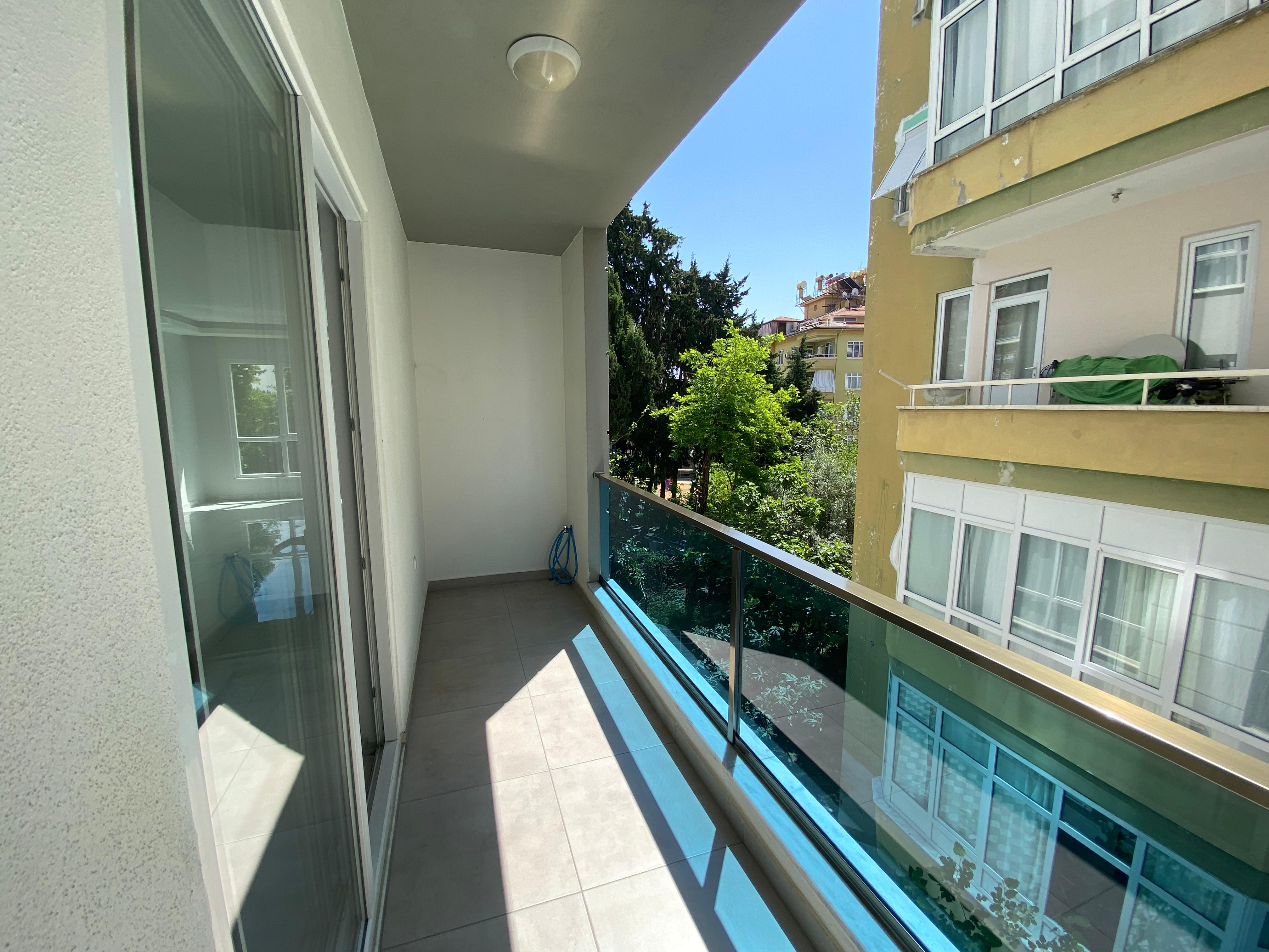 Satılık 2+1 dairenin kapalı balkonunda cam korkuluk, sürgülü kapı ve ağaçların arasındaki cephe