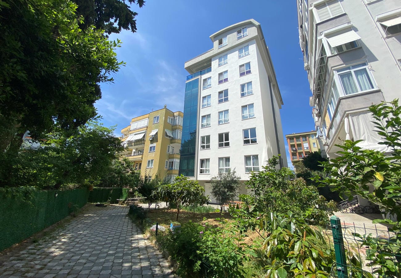 Alanya’da satılık 2+1 dairenin çevresinde mavi camlı cephe, bahçe yolu ve meyve ağaçları