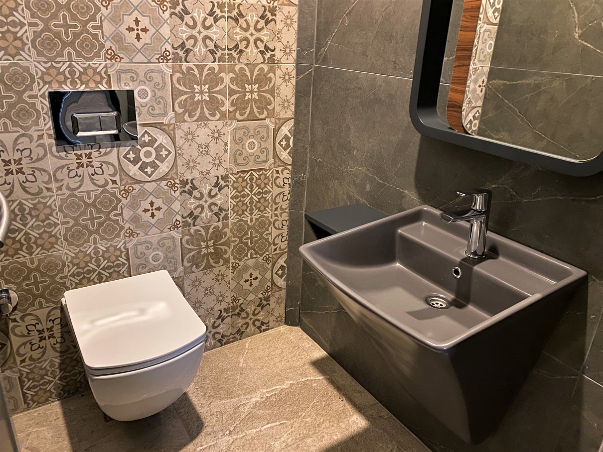 Banyoda asma koyu gri lavabo, desenli duvar karosu ve siyah çerçeveli ayna daire içinde