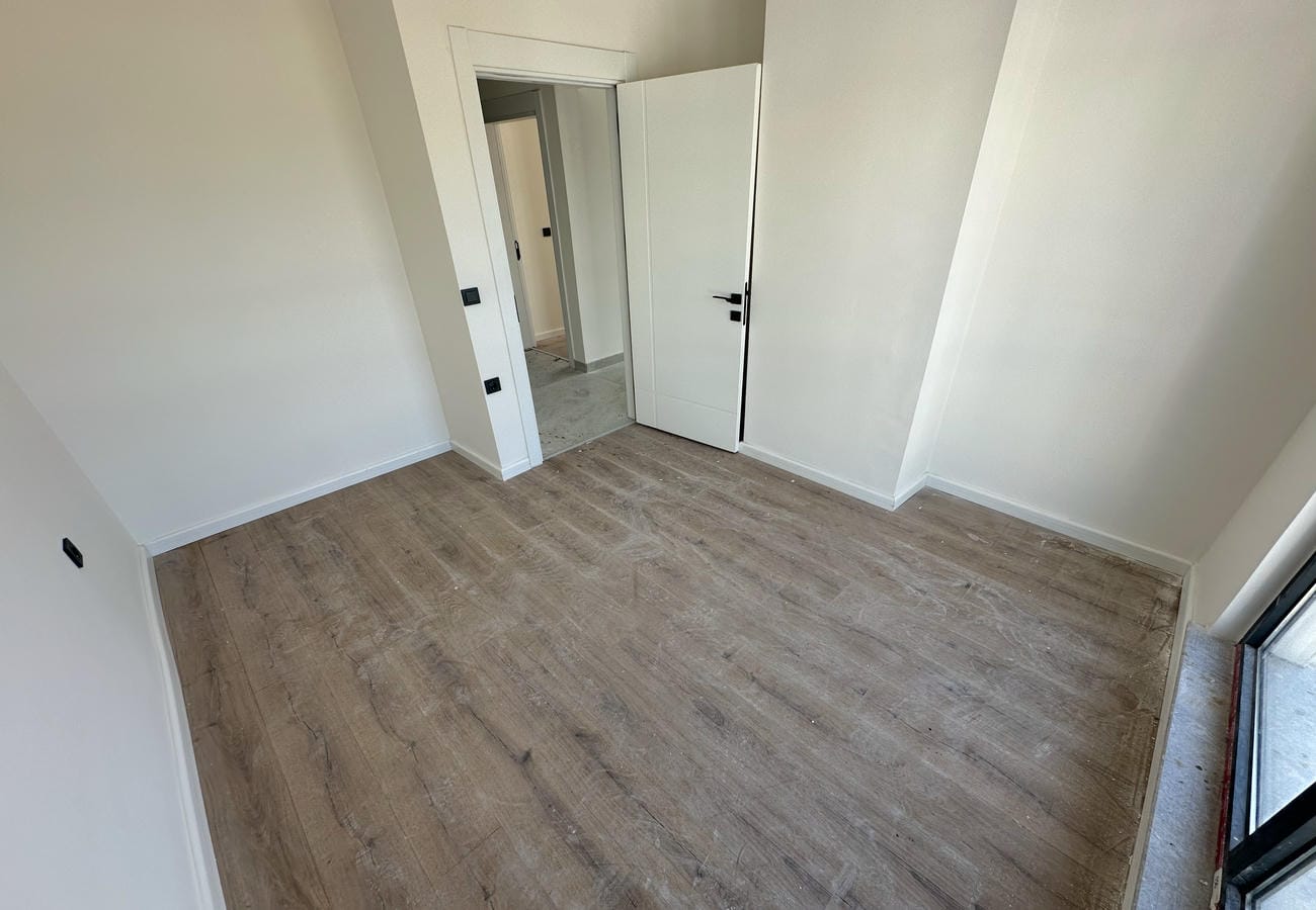 Boş yatak odasında panoramik balkon kapısı, beyaz duvarlar ve açık iç kapı