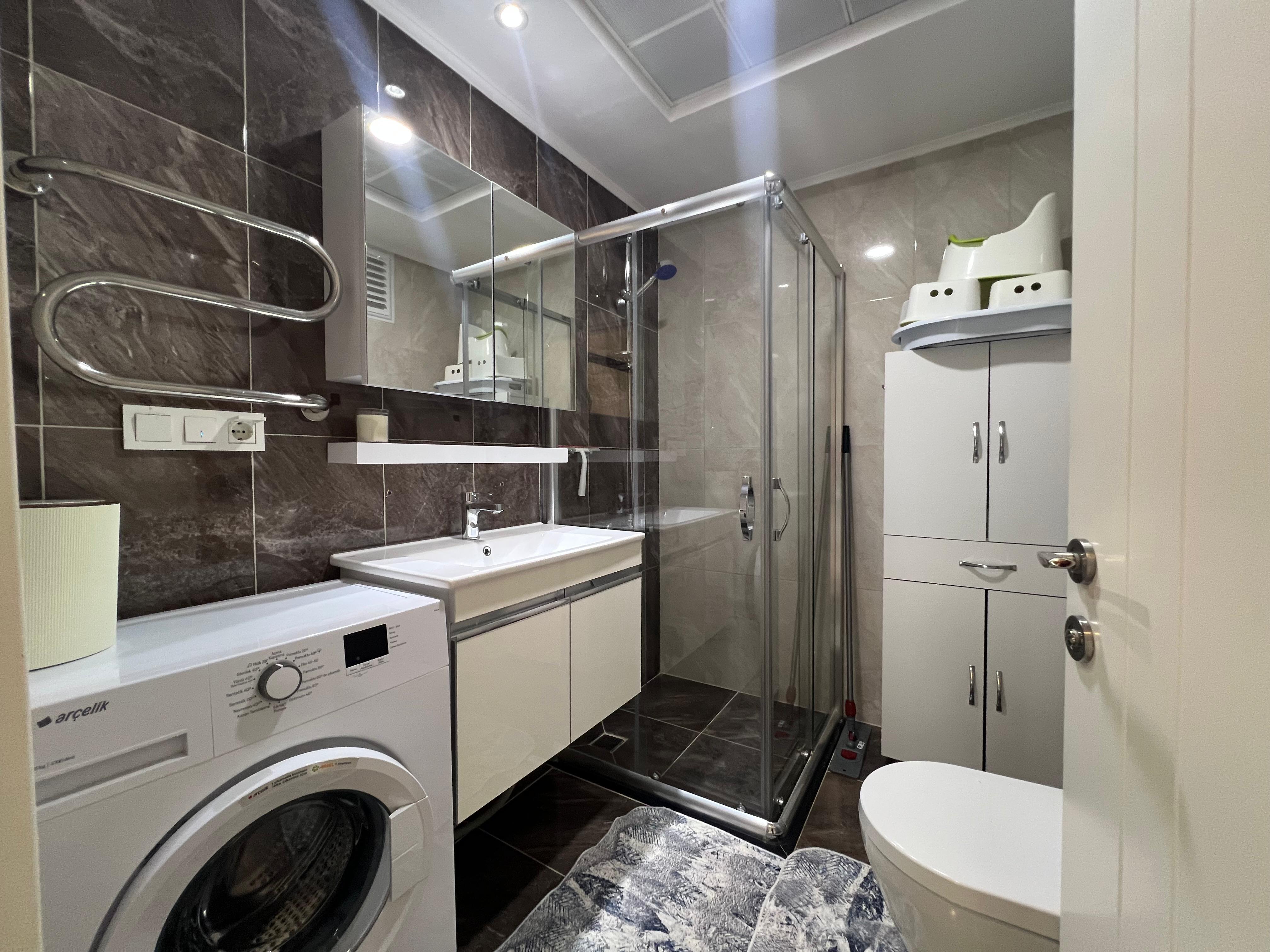 Bathroom with a glass shower cabin washing machine and dark marble-patterned tiles in an apartment in Avsallar