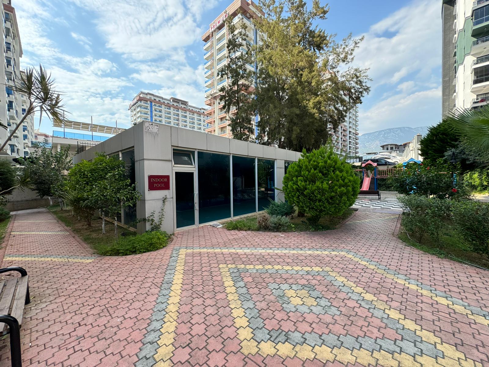 Dairelerin bulunduğu rezidansın bahçesinde oyun alanı yanındaki cam cepheli tek katlı yapı