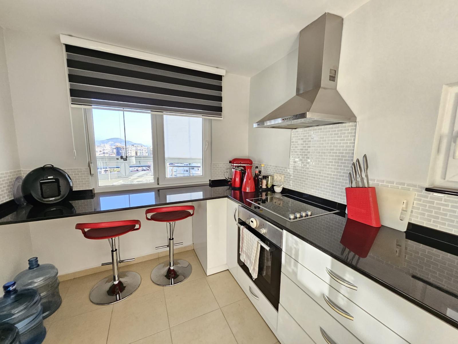 Siyah tezgâhlı köşe mutfakta beyaz dolaplar ve iki kırmızı bar sandalyesi bulunan penthouse