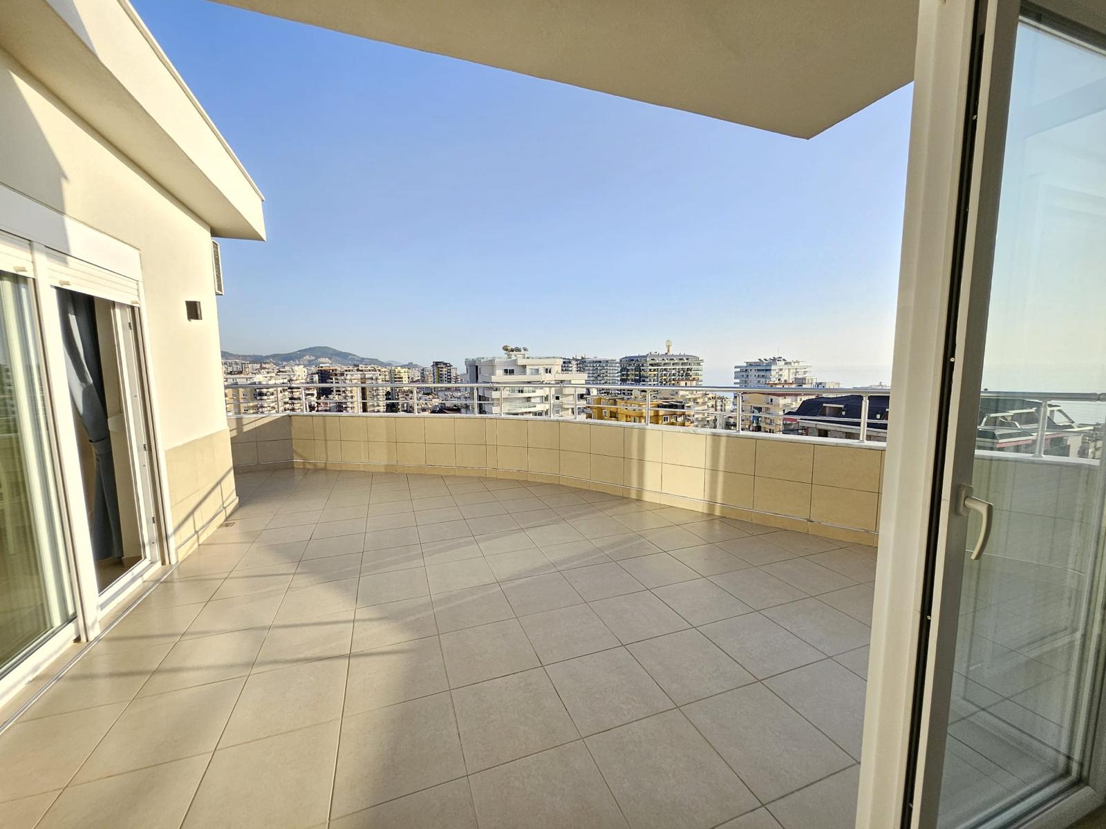 Penthouse yanında geniş açık terasta metal korkuluk, cam kapılar ve dağ manzaralı şehir panoraması