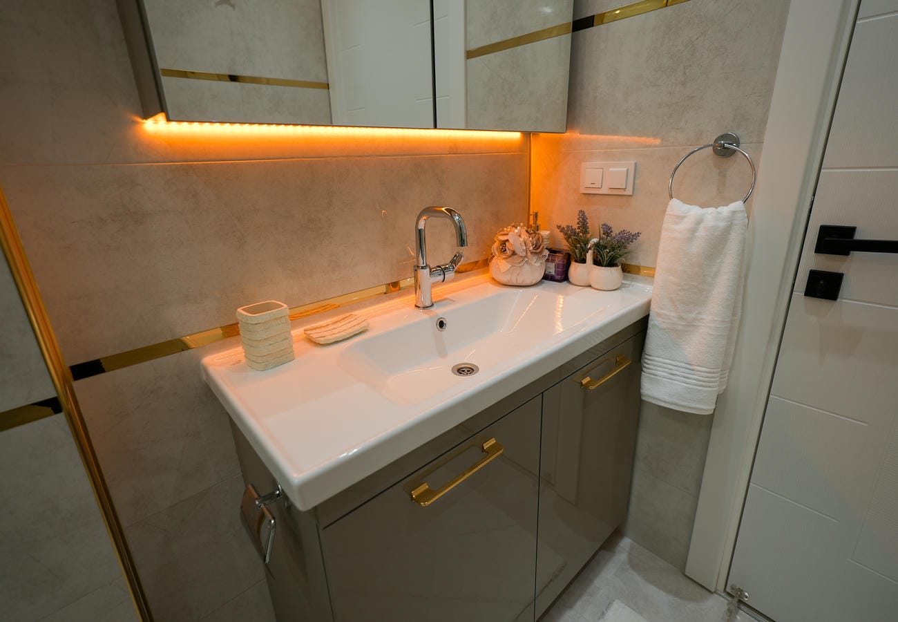 Mahmutlar'daki penthouse banyosunda dikdörtgen beyaz lavabo, gri dolap ve altın renk şeritli fayans