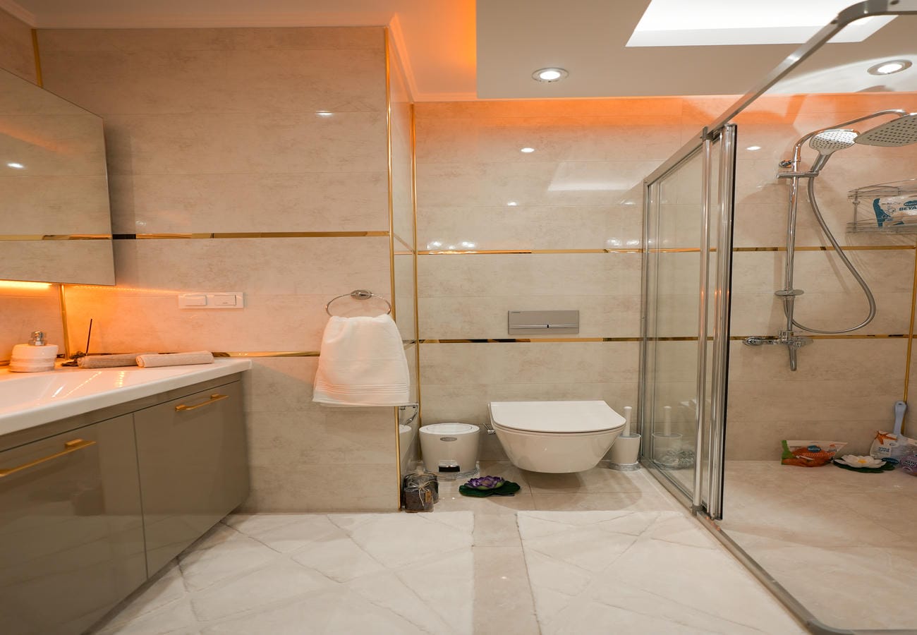 Alanya'da geniş cam duşakabinli banyoda beyaz lavabo, altın renk duvar şeritleri ve tavan aydınlatması