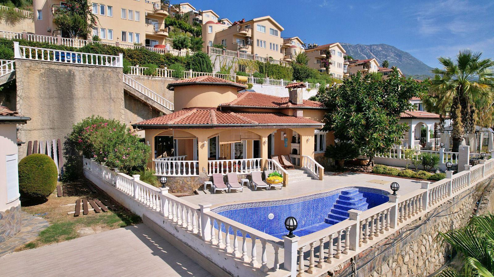 A villa for sale has an open pool, covered terrace and red tile roof among palms