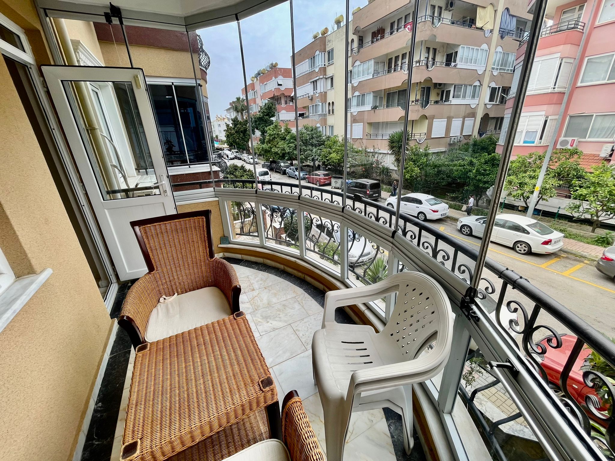 Dairenin balkonunda beyaz minderli hasır koltuk, açık kapı ve sokaktaki park edilmiş araçlar