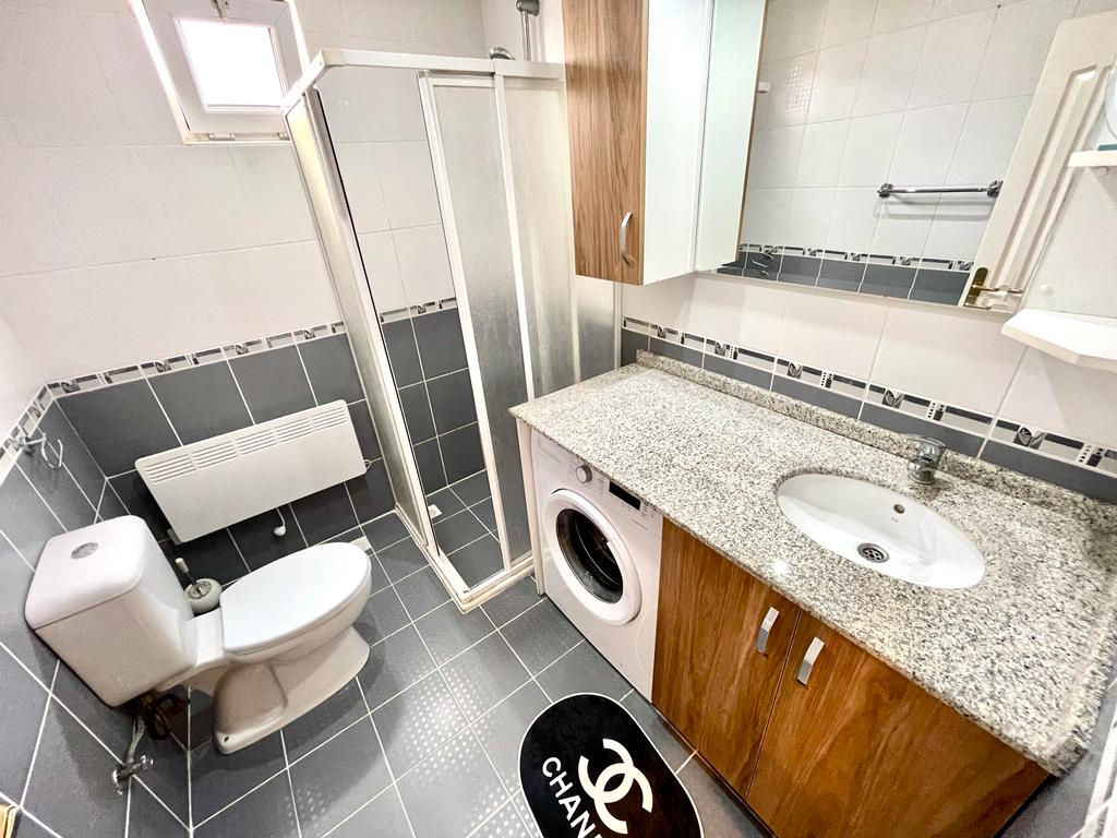 Banyoda buzlu cam duş kabini, granit tezgâhta oval lavabo ve çamaşır makinesi