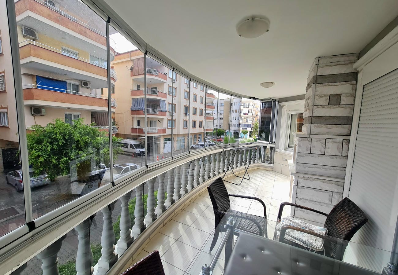 Dairenin camla kapatılmış balkonunda cam masa, siyah örgü koltuklar ve konutlara bakan manzara