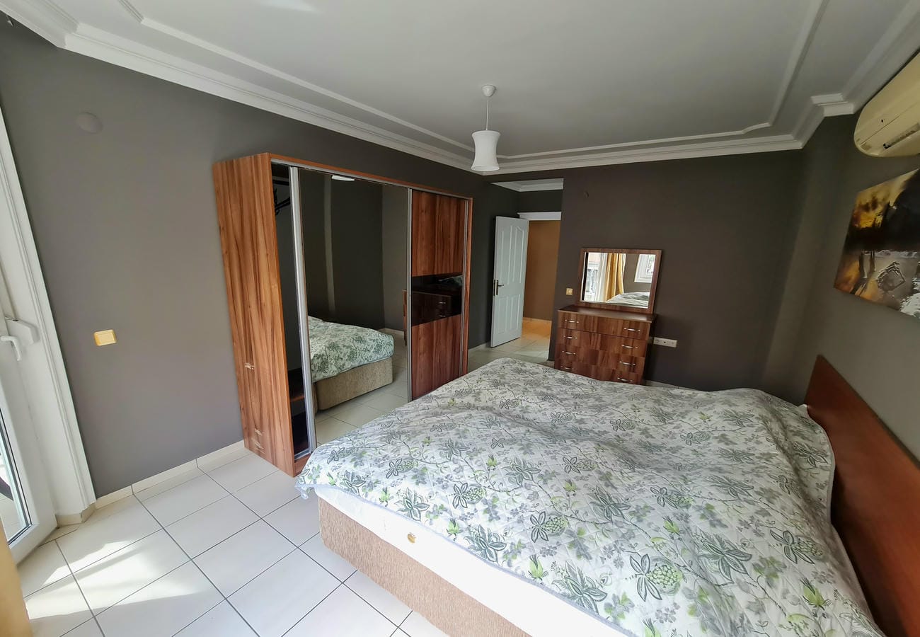 Yatak odasında 2+1 konuta ait desenli örtülü çift kişilik yatak, aynalı şifonyer ve cam balkon kapısı