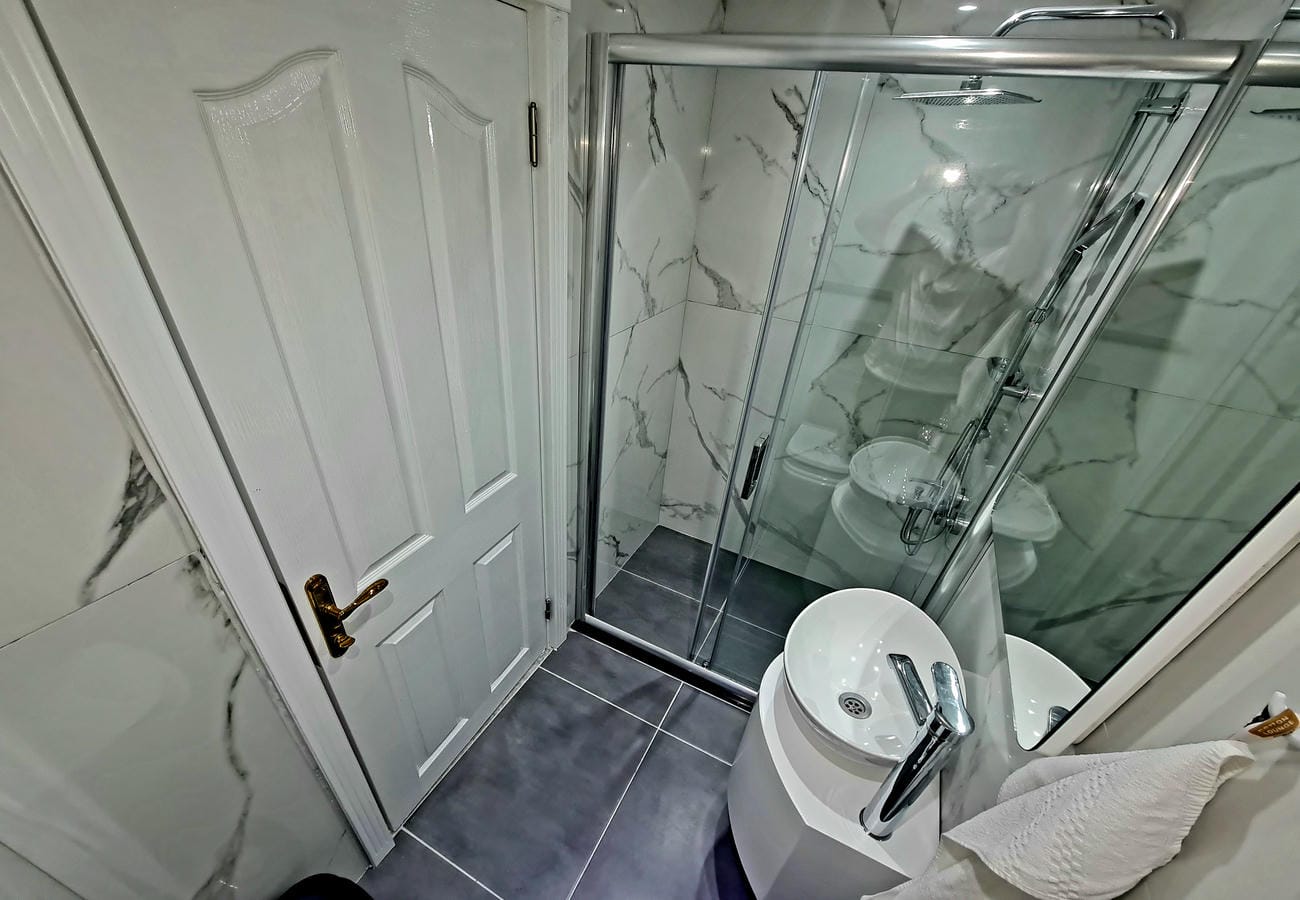 Dairenin banyosunda cam duş kabini, beyaz lavabo dolabı ve beyaz mermer görünümlü duvar fayansı
