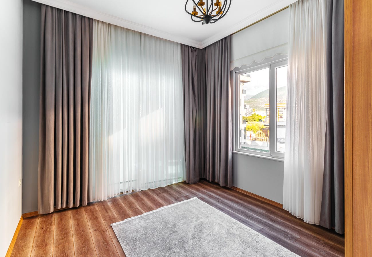 Boş yatak odasında gri duvarlar, köşe pencere ve perdelerin ardında balkon çıkışı