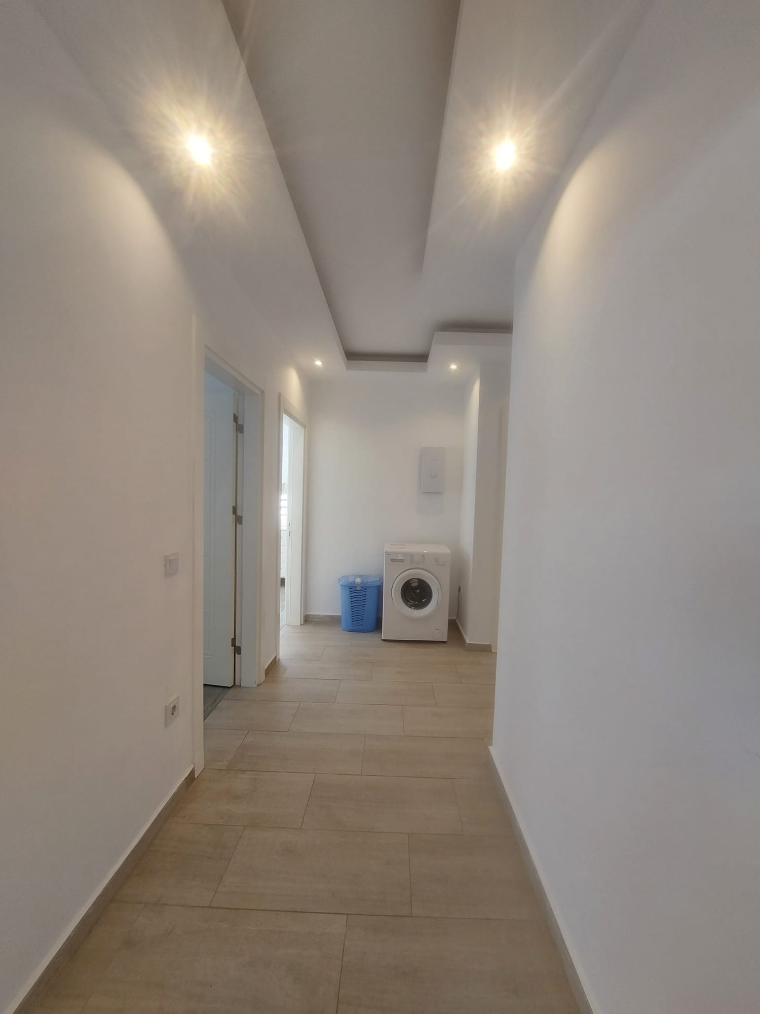 Uzun aydınlık koridorda sol tarafta kapı girişleri ve ileride çamaşır makinesi bulunuyor