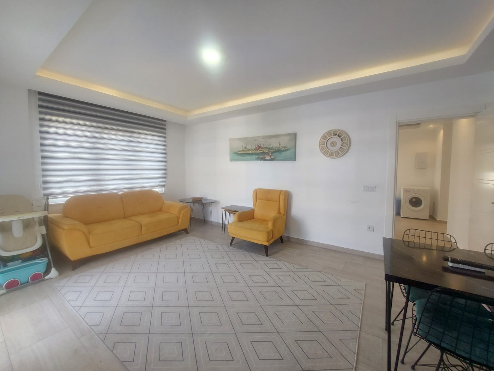 Oturma odasında sarı kanepe ve koltuk geometrik desenli halının yanında 2+1 konutta yer alıyor