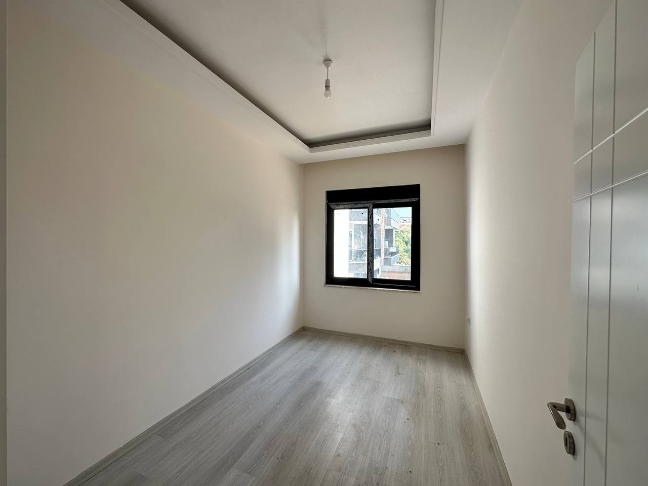 Dar boş yatak odasında siyah çerçeveli pencere, açık laminat zemin ve komşu binalara bakış