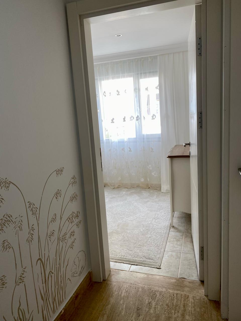 Dairenin yatak odasında beyaz kapı ardından büyük pencere, şifonyer ve desenli halı