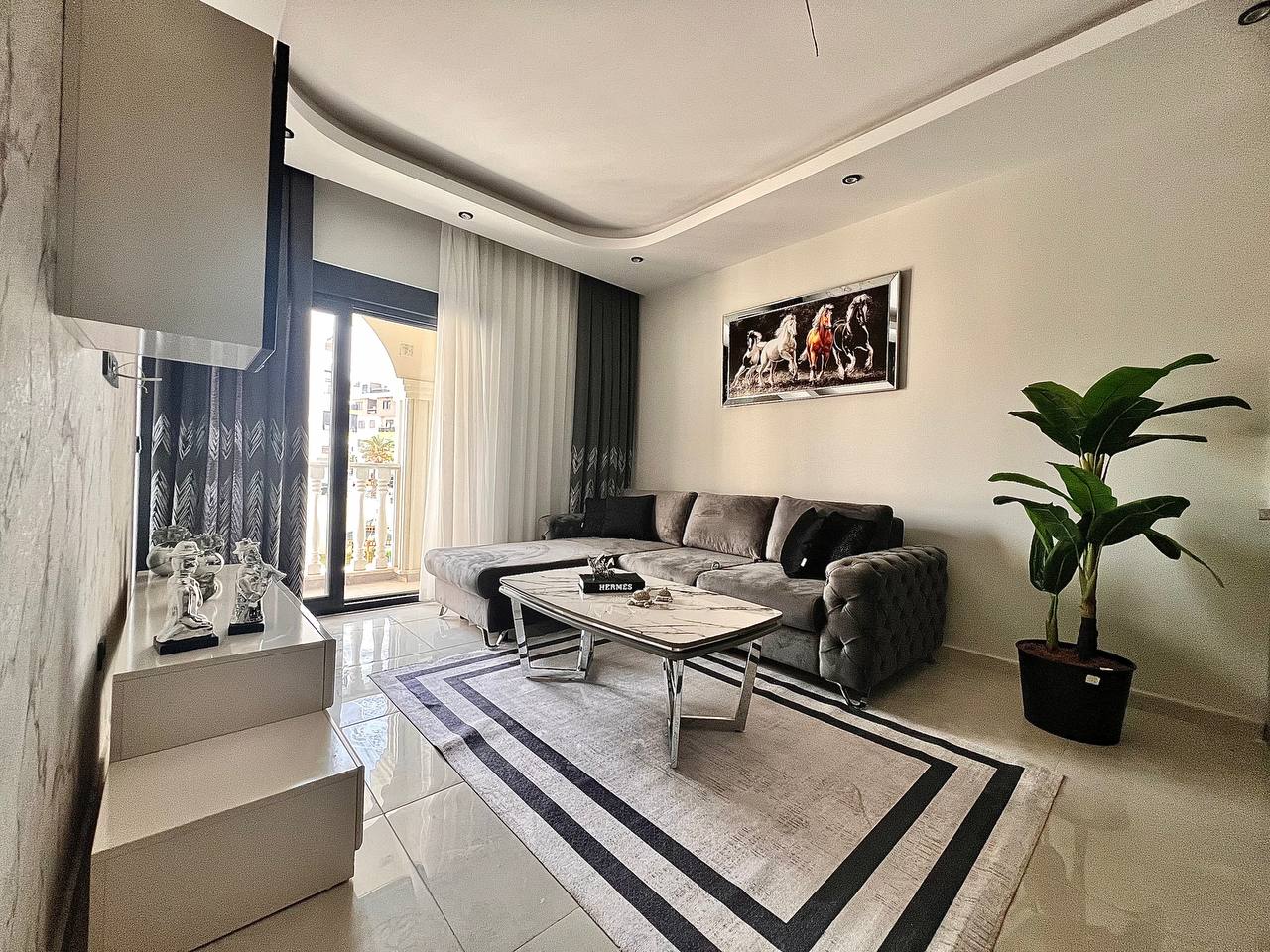 1+1 dairenin mutfaklı salonunda gri köşe koltuk, mermer sehpa ve balkon kapısı