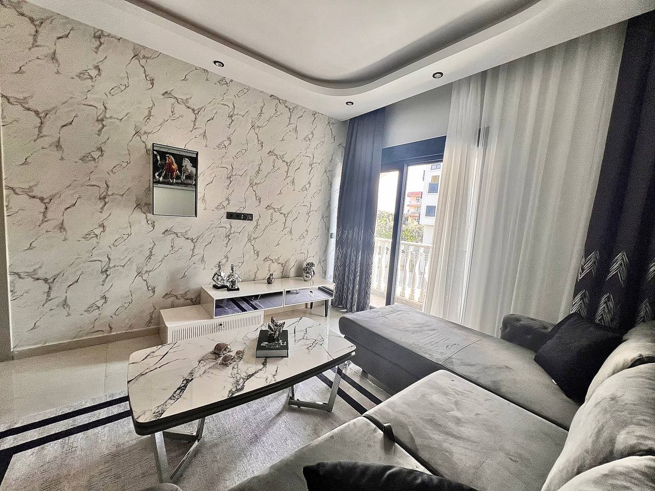 Salonda gri köşe koltuk, mermer desenli sehpa, duvar aynası ve perdeli balkon kapısı