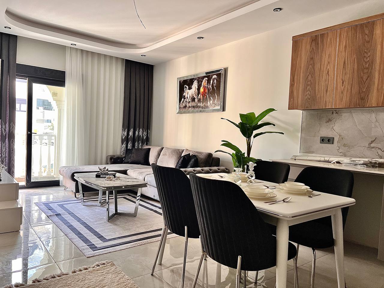 Oba’daki 1+1 dairenin mutfaklı salonunda köşe mutfak, servisli masa ve balkon kapısı