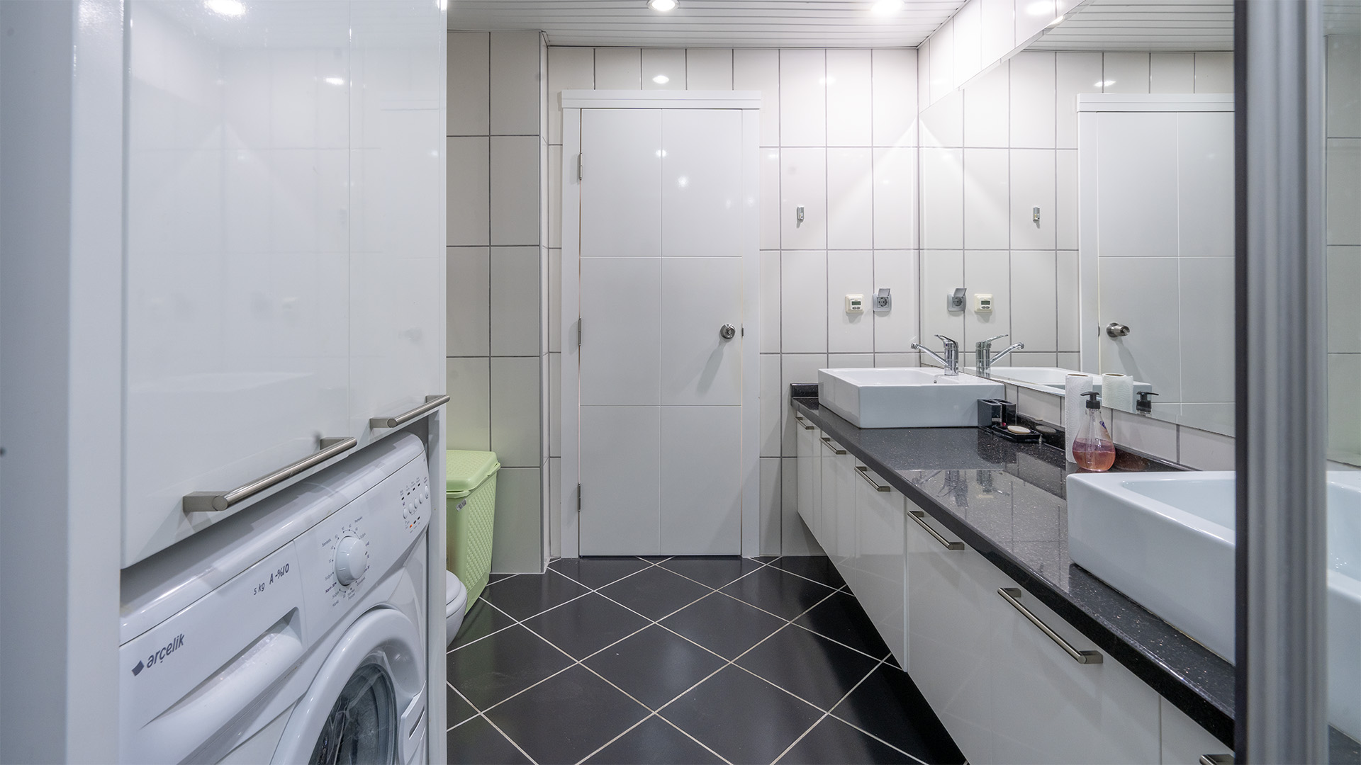 Dairenin banyosunda iki beyaz lavabo, gri tezgâhlı uzun dolap ve büyük duvar aynası