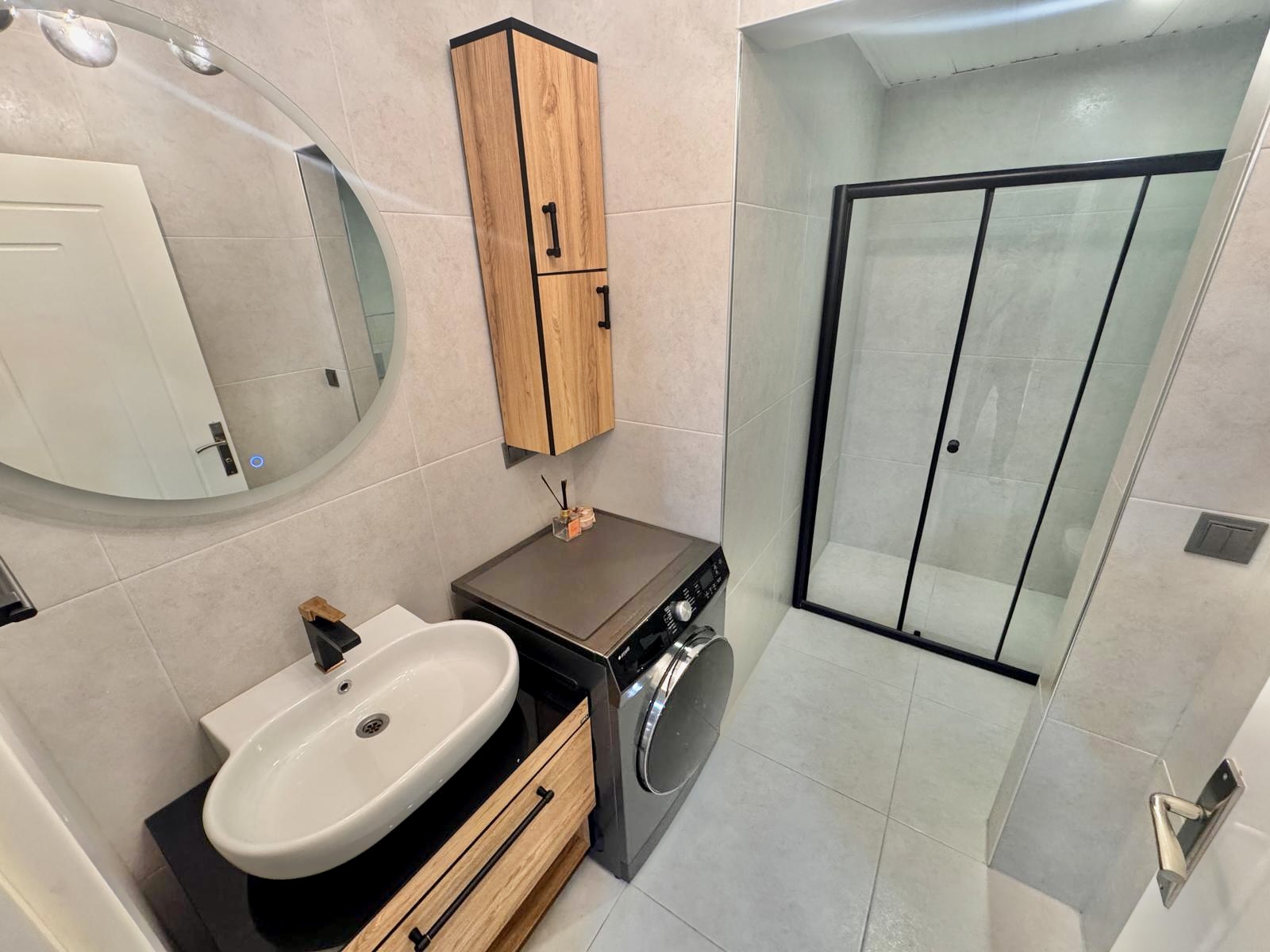 Siyah profilli duş alanı olan banyoda oval beyaz lavabo, çamaşır makinesi ve ahşap dolap