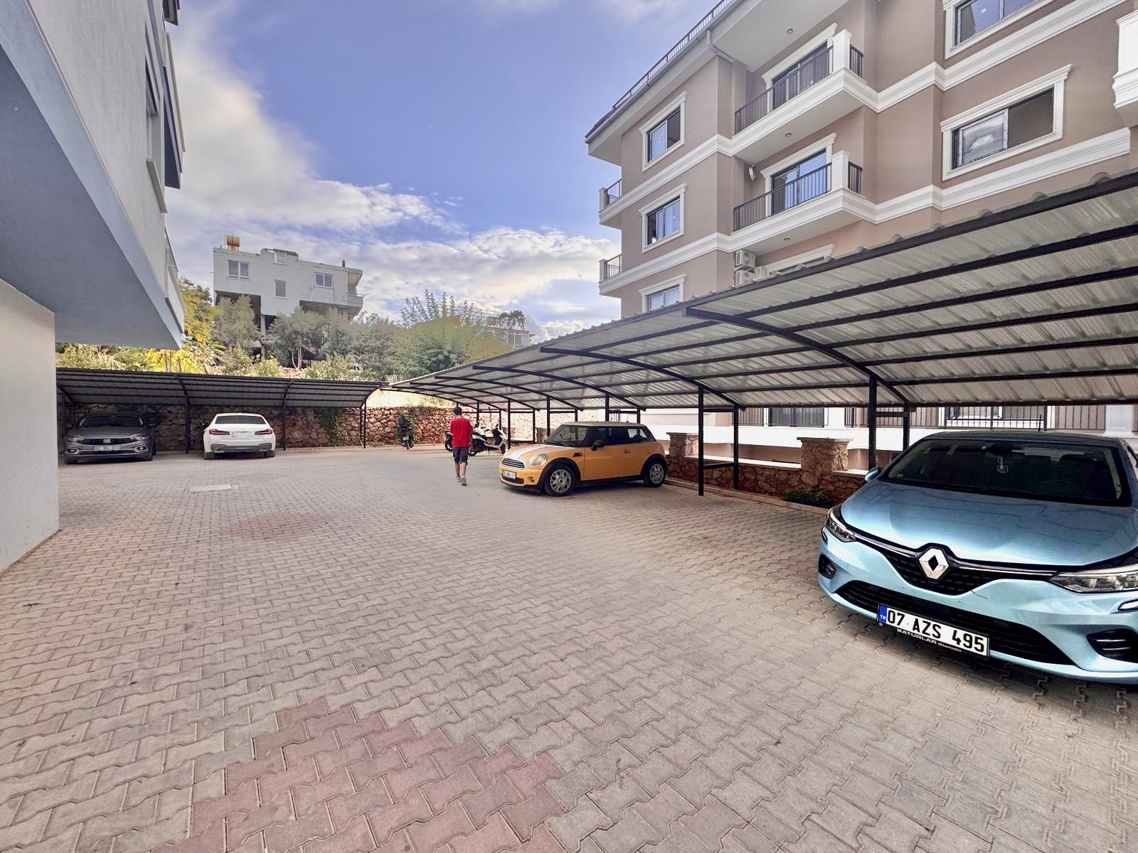 Gri parke taşlı otoparkta metal sundurma altında sarı araç, önde mavi araç ve balkonlu cephe