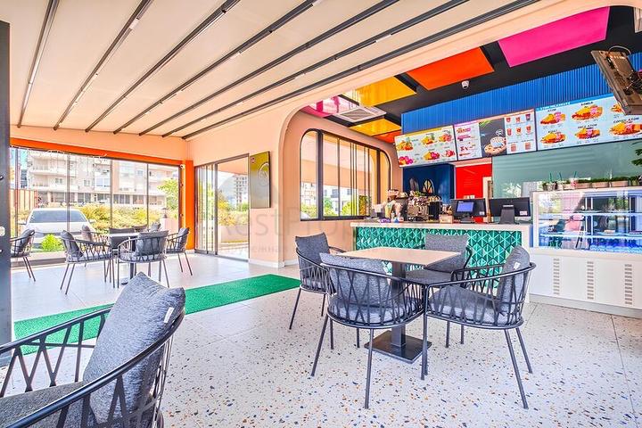 Cafe with a turquoise tiled counter, woven armchairs, and bright ceiling panels by glass doors