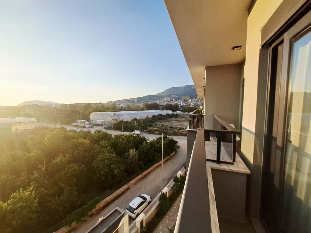 Long balcony with clear railing and evening views of the road, garden, and mountains at a flat