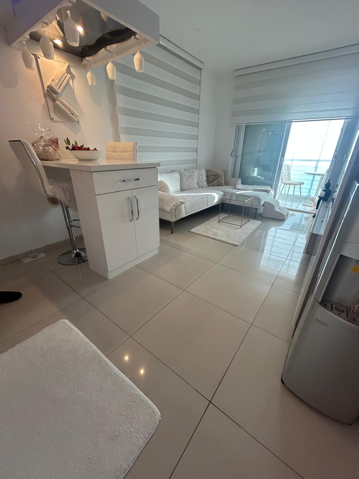 Dairedeki mutfak-salonda beyaz köşe koltuk ve bar tezgâhı, panoramik balkon camlarıyla