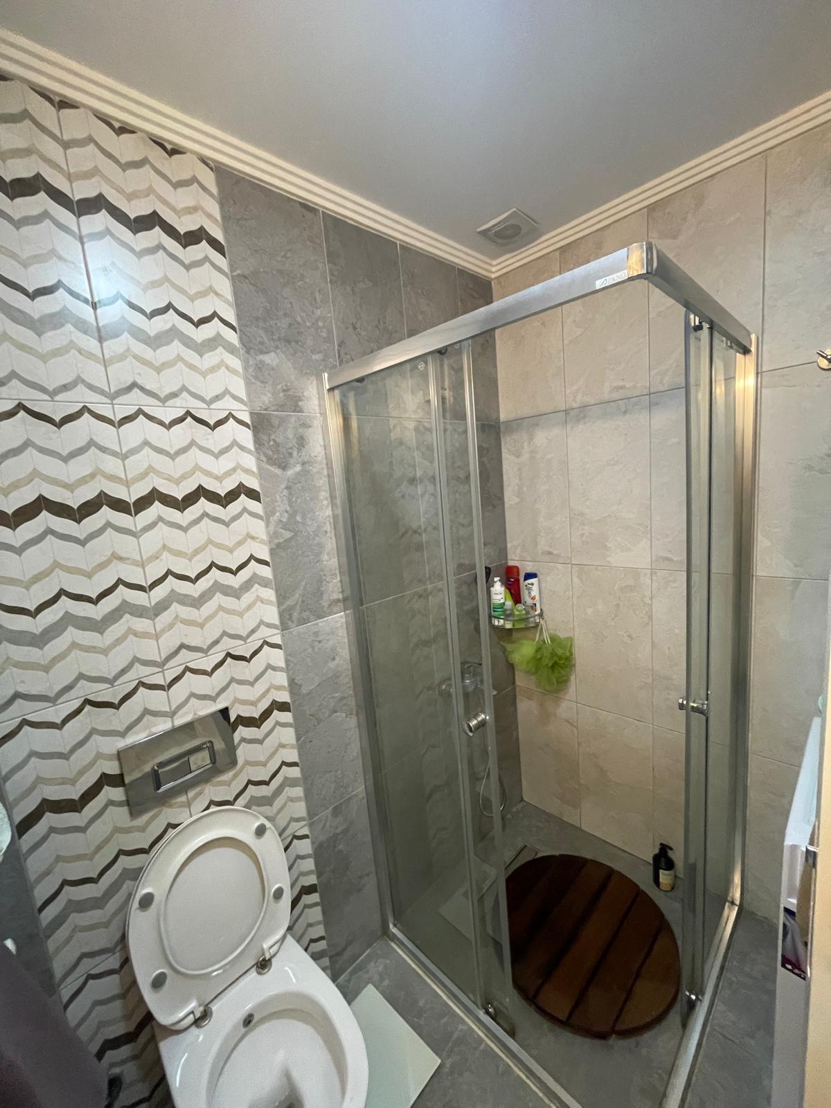 Cam duş kabinli banyo, askılı klozet ve ahşap duş paspaslı konutta