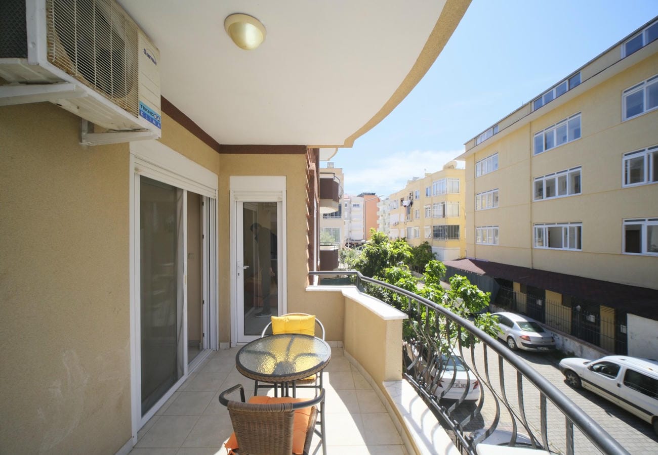 Kavisli cam balkon, yuvarlak cam masa ve hasır sandalyeleriyle karşıdaki çok katlı konutlara bakıyor