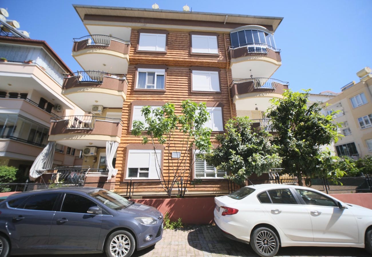 Kahverengi ahşap detaylı konut cephesinde yuvarlak balkonlar ve önünde yeşil ağaçlar bulunan bir daire görülüyor