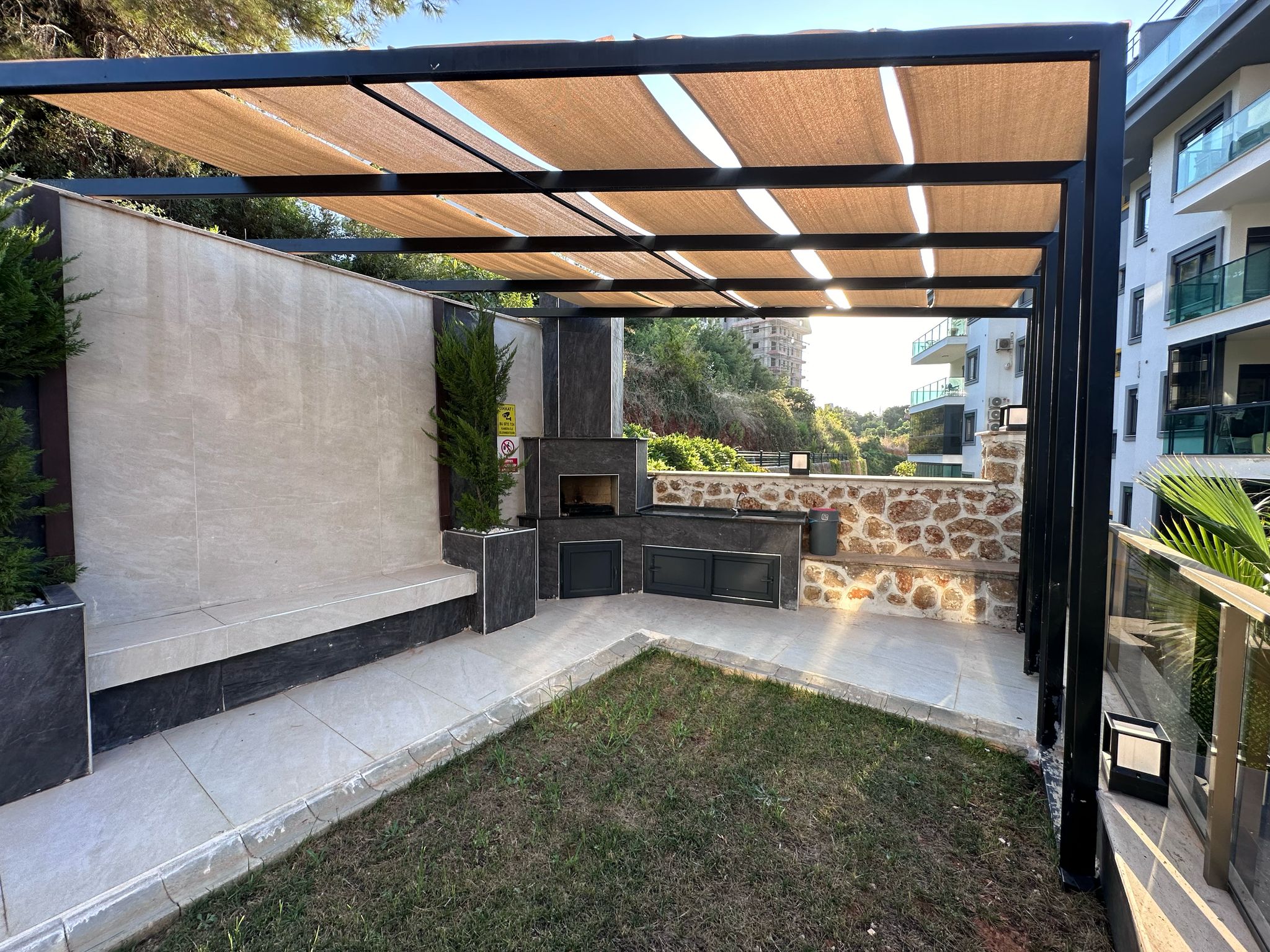 Kumaş tenteli metal pergola, çim alanı ve taş duvarıyla Oba’daki dairelerin bulunduğu rezidansta