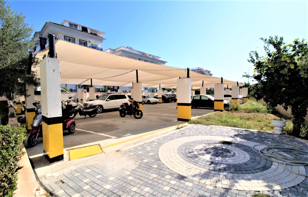 Kapalı otoparkta otomobiller, motosikletler ve sarı siyah direkli tente yapısı dairelerin bulunduğu komplekste