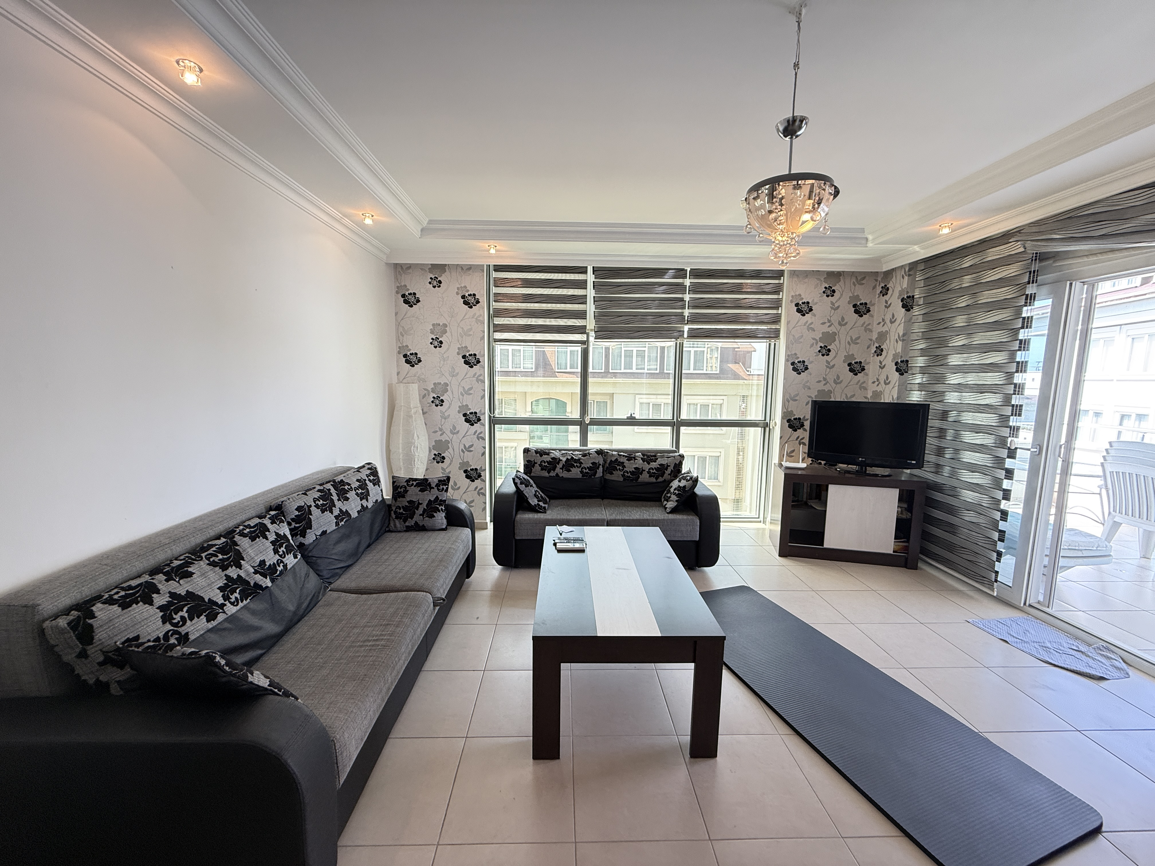 Panoramik camlı salonda gri koltuk, televizyon ve balkon kapısı