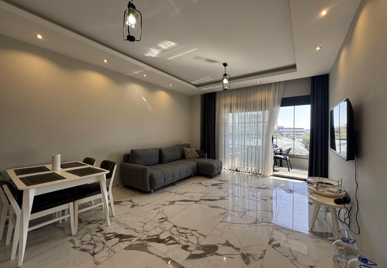 Gri köşe koltuklu salon satılık konutta televizyon ve panoramik balkon kapısıyla düzenlenmiş