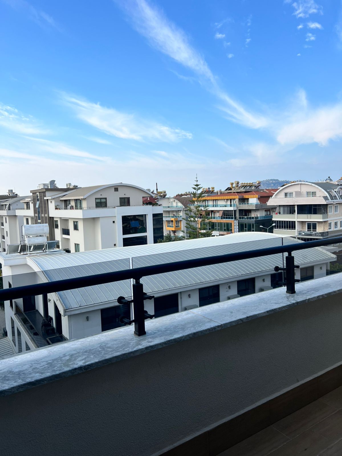 Geniş beton parapetli cam korkuluklu balkon, komşu binalar ve bulutlu gökyüzüyle şehir dokusuna bakıyor