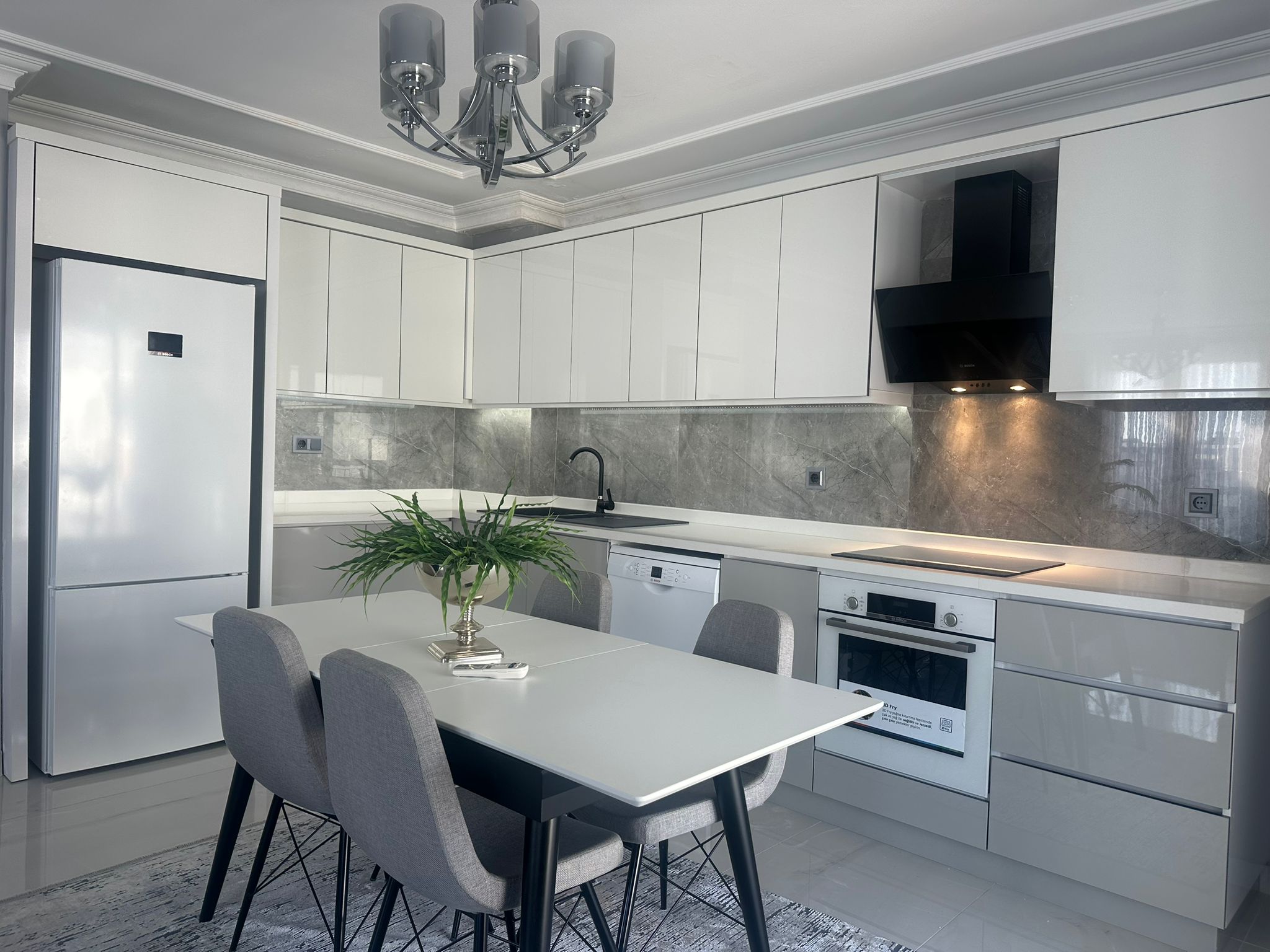 Parlak beyaz dolaplı ve gri taş tezgâhlı mutfak Oba’daki penthouse dairede yemek alanına açılıyor