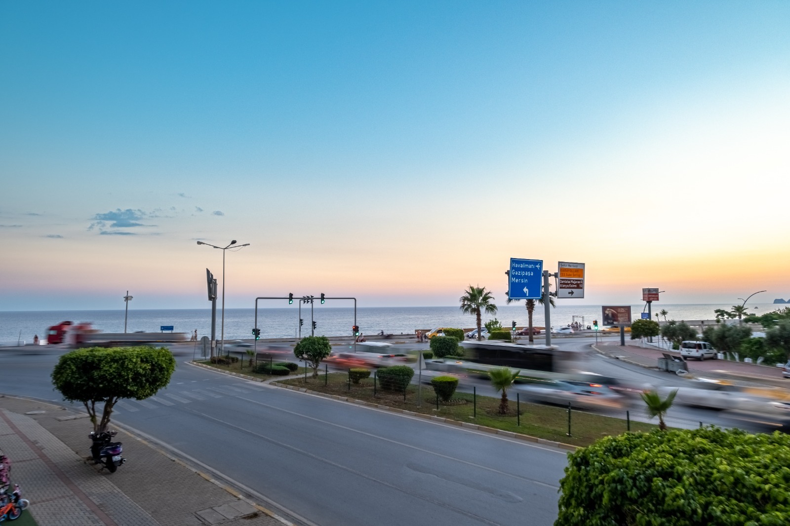 Akşam saatlerinde kavşak sonrası denize uzanan sahil yolu, palmiyeler ve trafik ışıkları